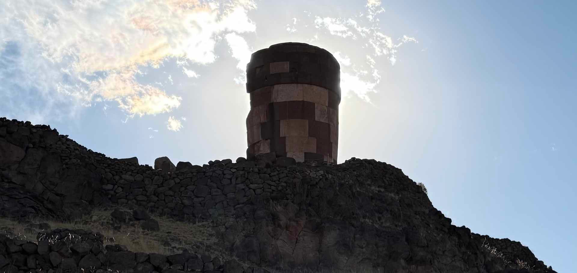 Cylindrical stone burial tower silhouetted against a bright sky with scattered clouds.