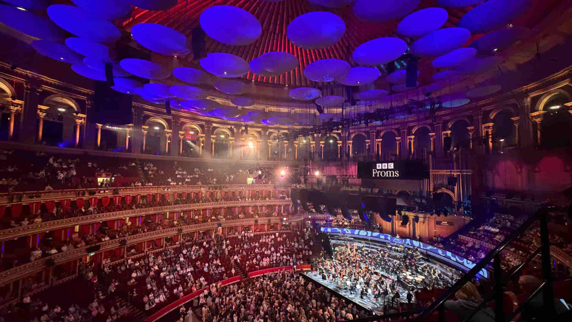 Elevated angle of the massive auditorium filled with classical music listeners beneath blue stage lighting