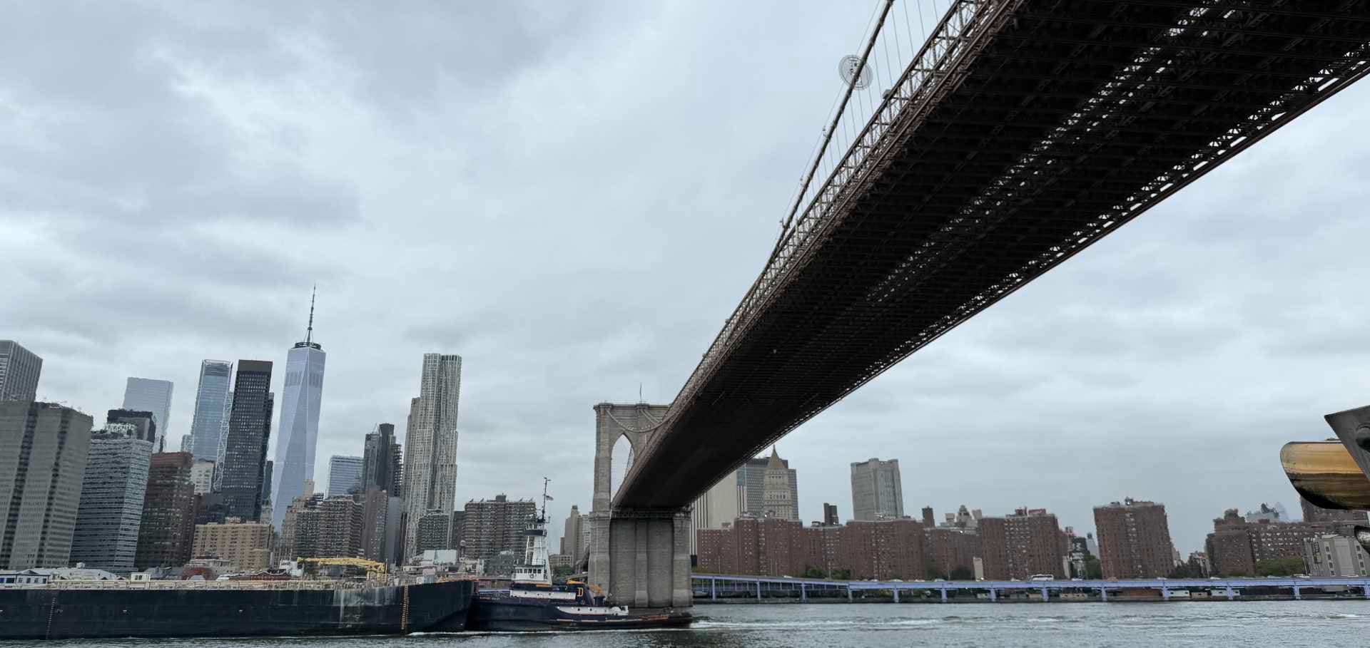 A tugboat navigates under the grand stone arches of the Brooklyn Bridge with the Manhattan skyline behind.
