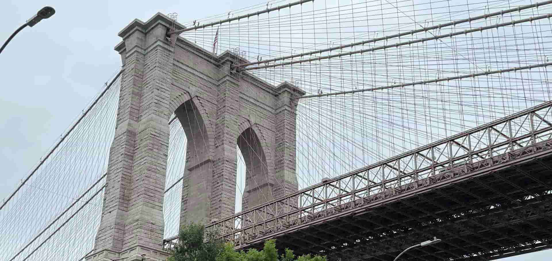 Side profile of the stone tower and structural iron framework of the Brooklyn Bridge against an overcast sky.