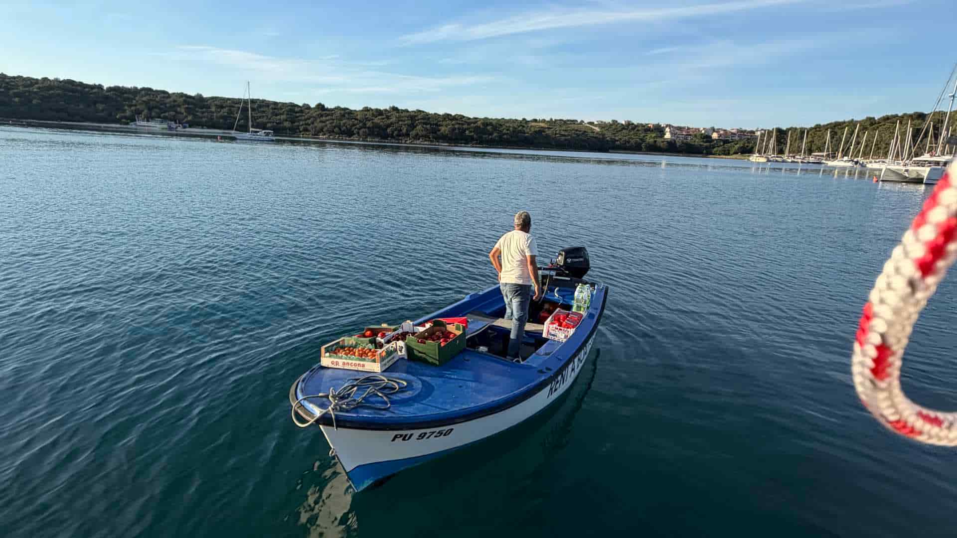 A small motorboat carrying fresh produce floats on the calm waters of Soline Bay near anchored yachts