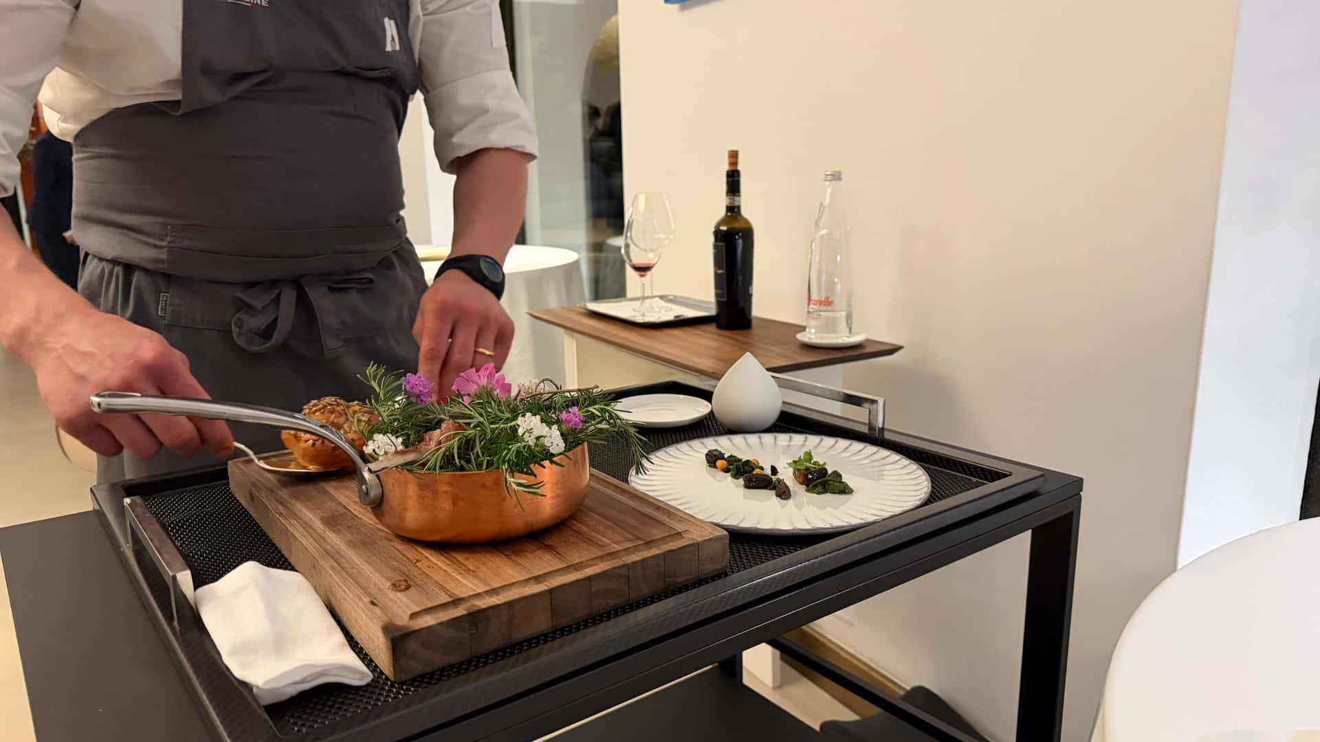 Chef plating delicate seafood and herbs from a copper pan on a serving cart.