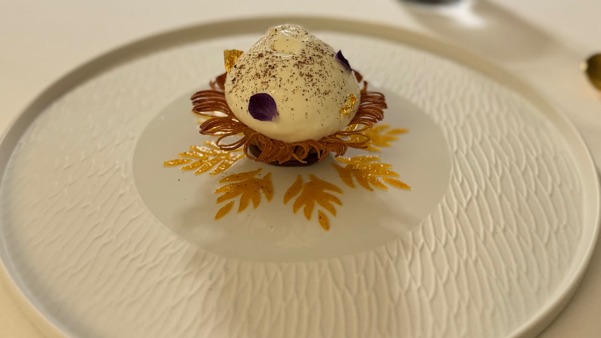 White dessert dome with gold leaf and floral details on a textured white plate.