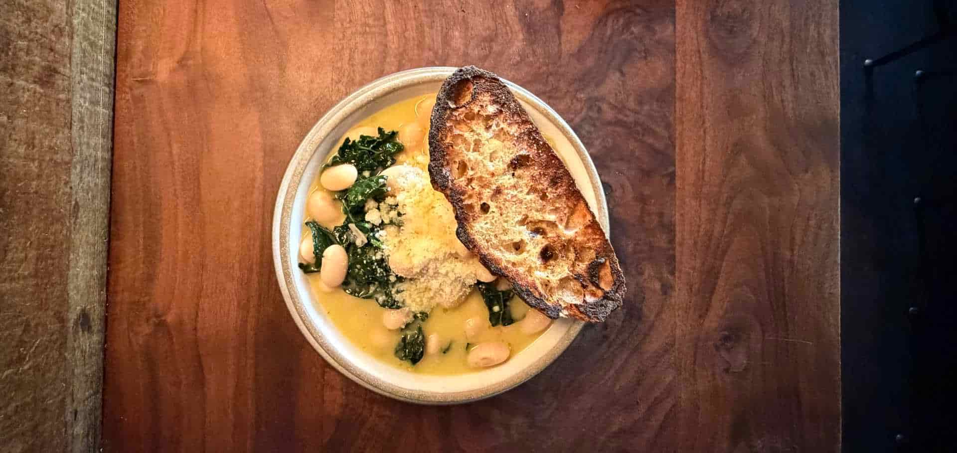 White bowl filled with white beans, greens, and a slice of toasted artisanal bread.